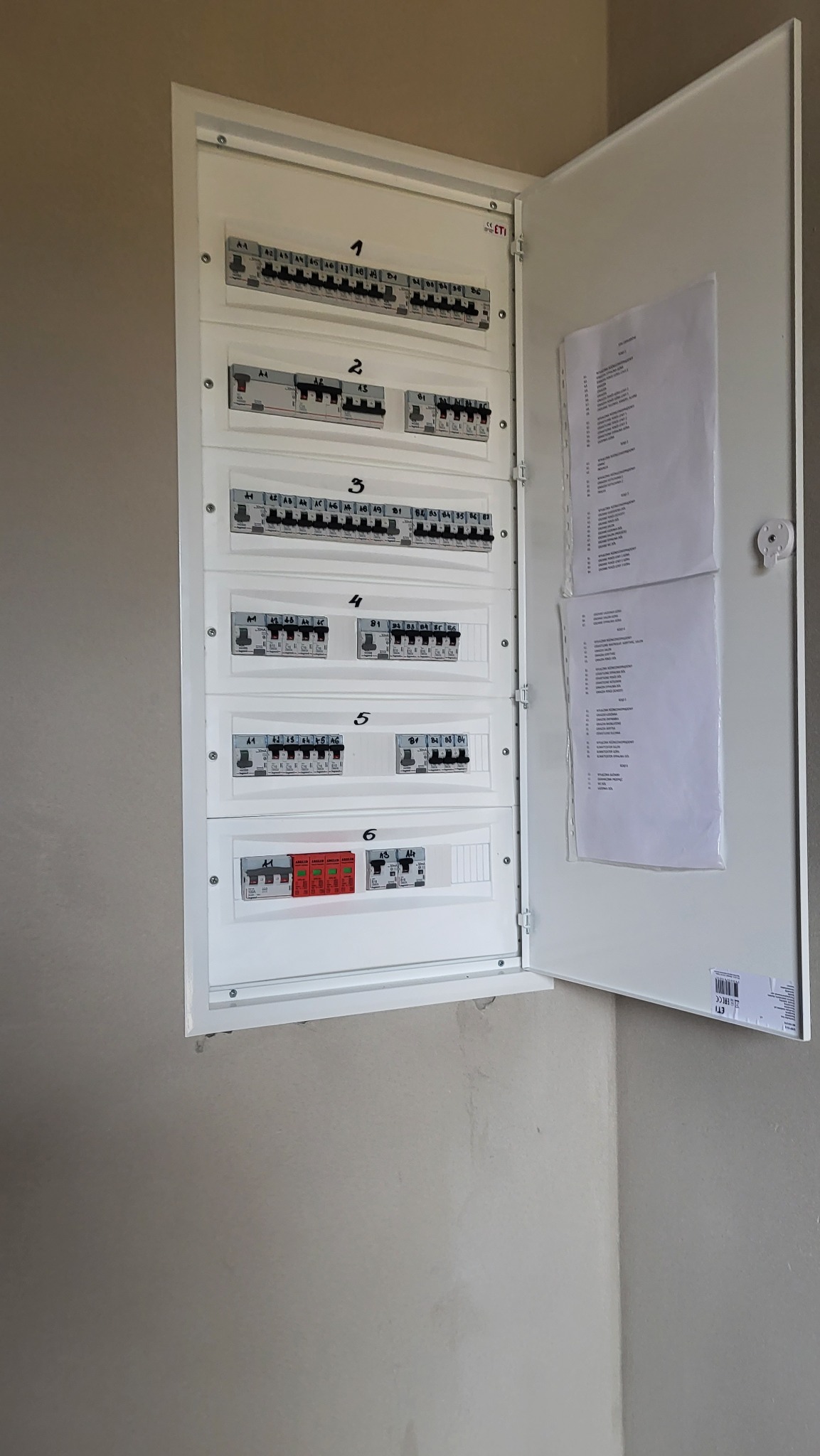 Otwarta skrzynka elektryczna z bezpiecznikami i wyłącznikami, ponumerowane sekcje, dokumentacja wewnątrz drzwiczek, widok z przodu na szarą ścianę.