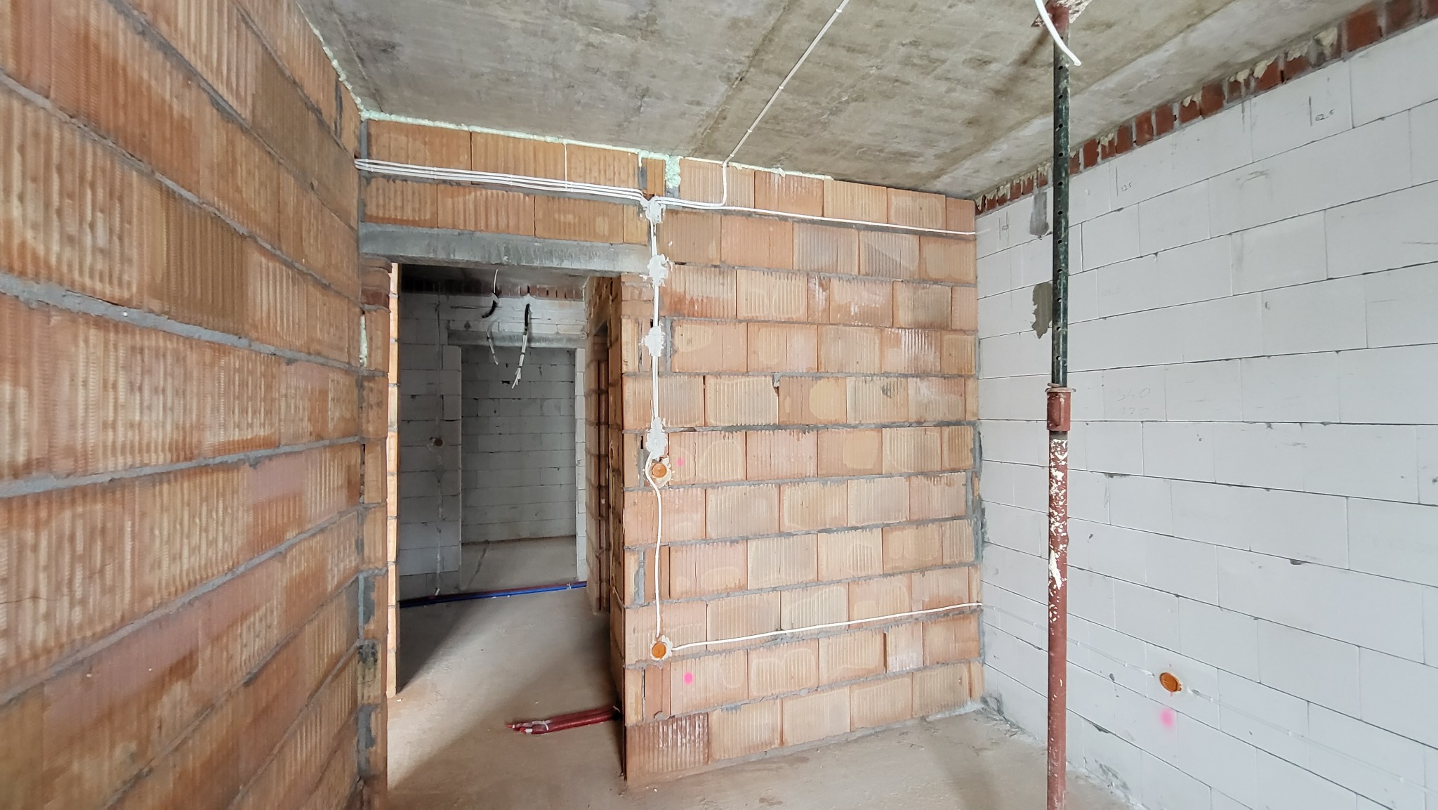 Surowy stan wnętrza budynku w trakcie prac elektrycznych: widoczne przewody elektryczne prowadzone po ścianach z cegły ceramicznej i bloczków, wspornik stalowy podtrzymujący strop.