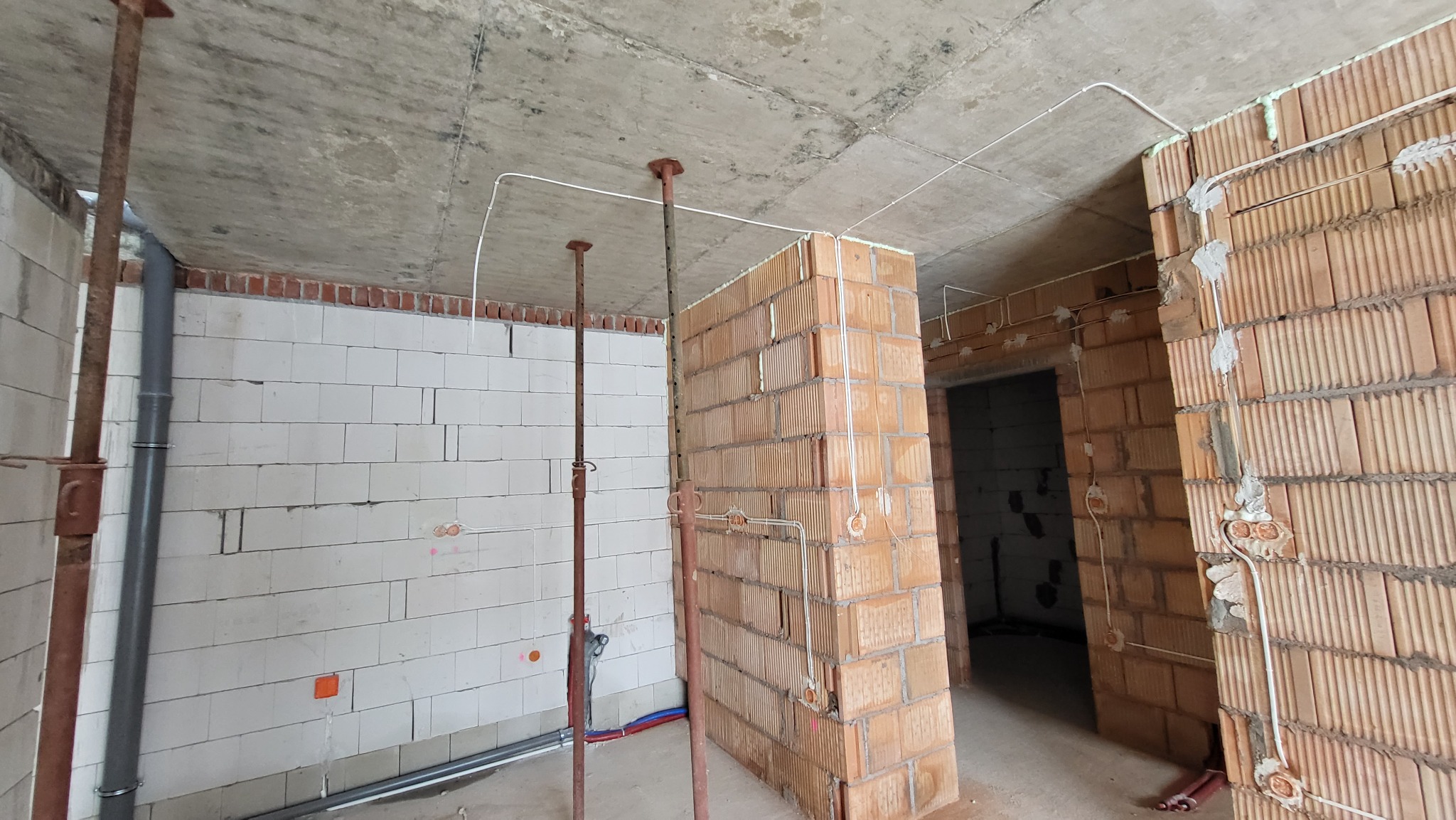 Surowy stan wnętrza w trakcie budowy: widoczne białe i czerwone bloki, betonowy sufit, stalowe podpory, wystające przewody elektryczne, puszki instalacyjne zamocowane na ścianach.