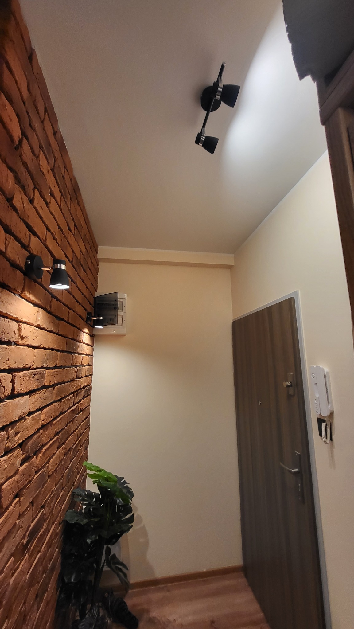 Korytarz z ceglaną ścianą, oświetleniem punktowym i skrzynką elektryczną, widok na drzwi wejściowe i domofon.