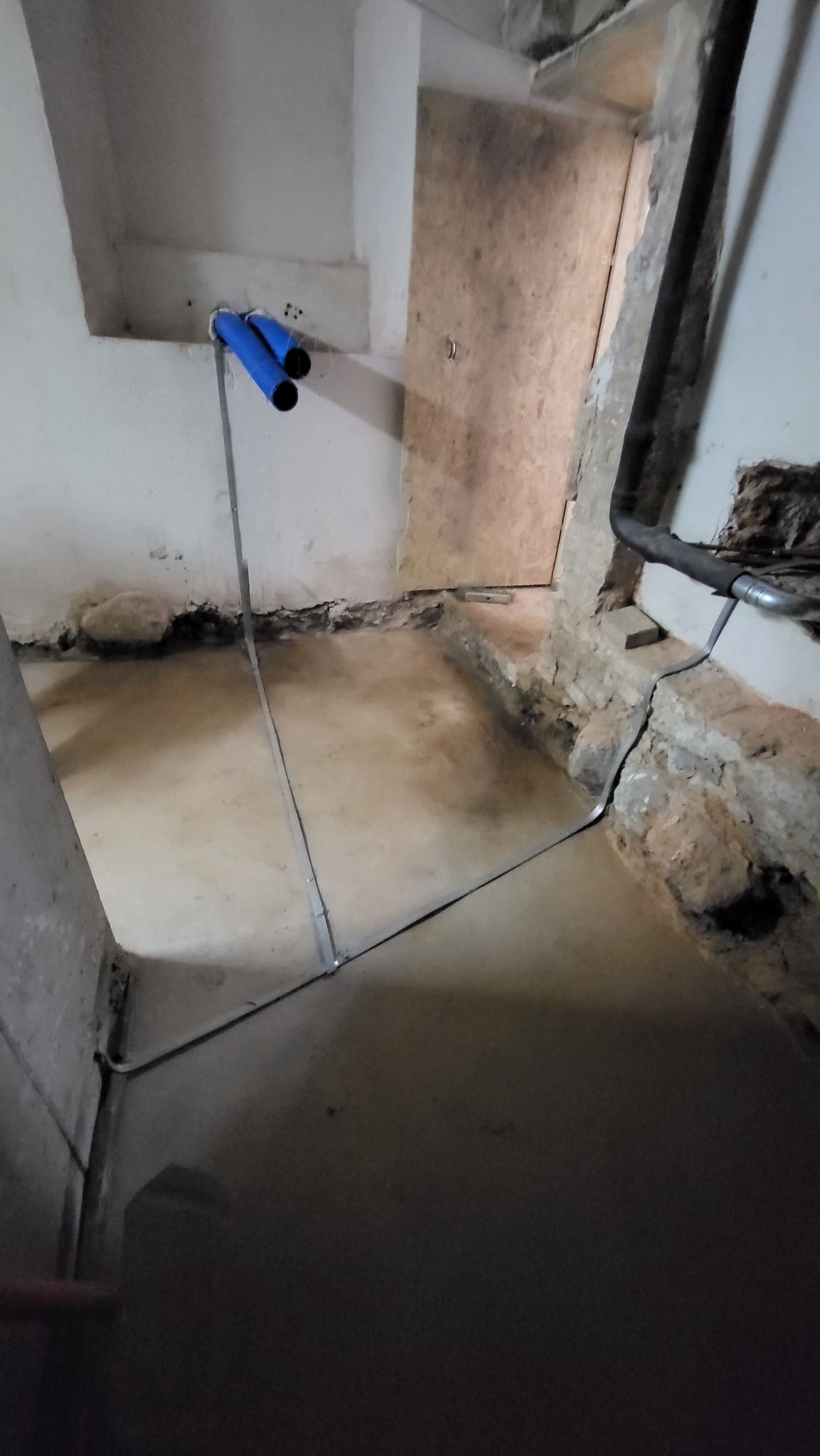 Niebieskie rury instalacji elektrycznej przymocowane do ściany w surowym pomieszczeniu z nieotynkowanymi ścianami i podłogą, widoczne przewody.