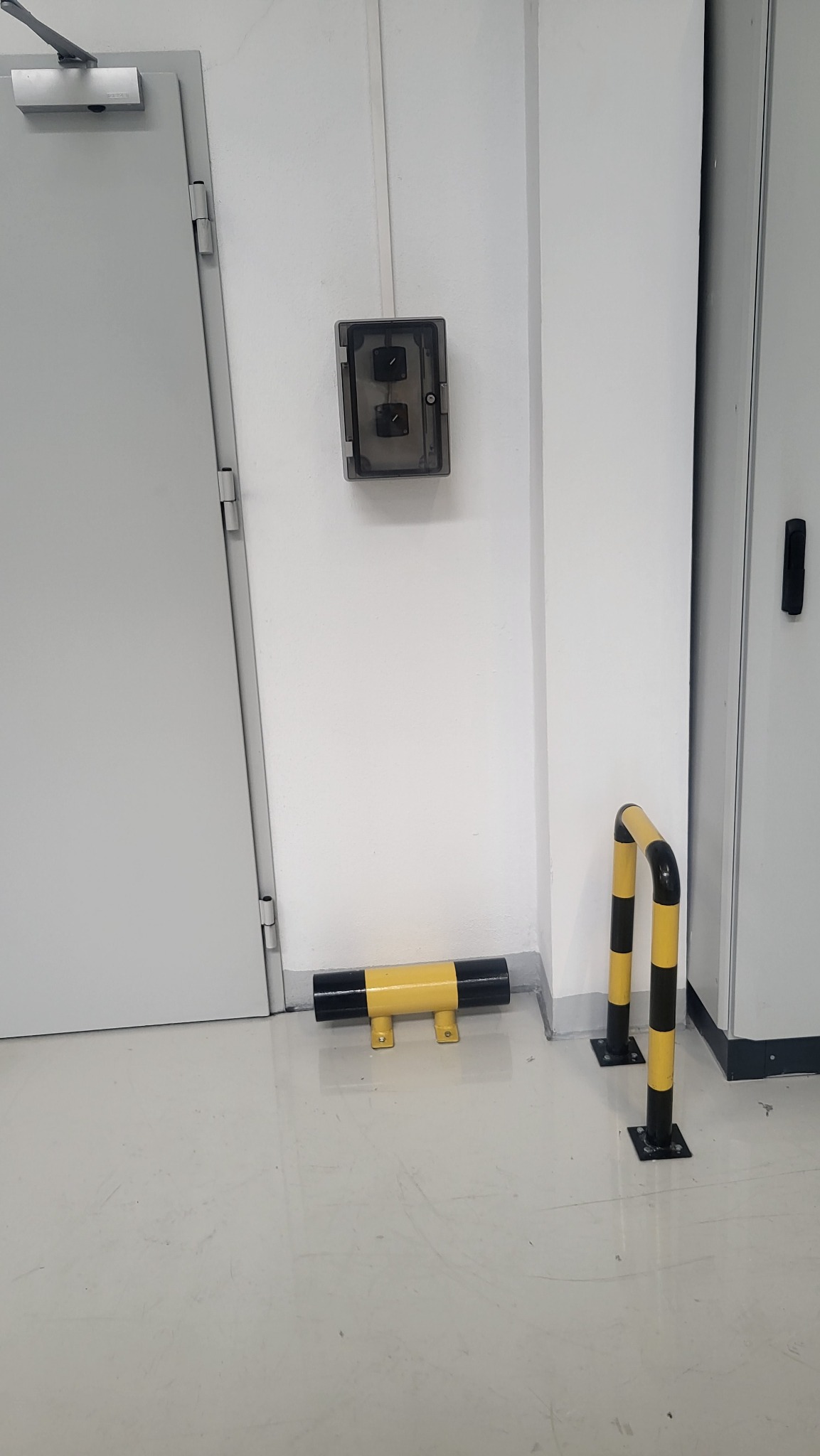 Szary włącznik dwuklawiszowy w przezroczystej obudowie na białej ścianie, obok drzwi i żółto-czarne odbojniki podłogowe.