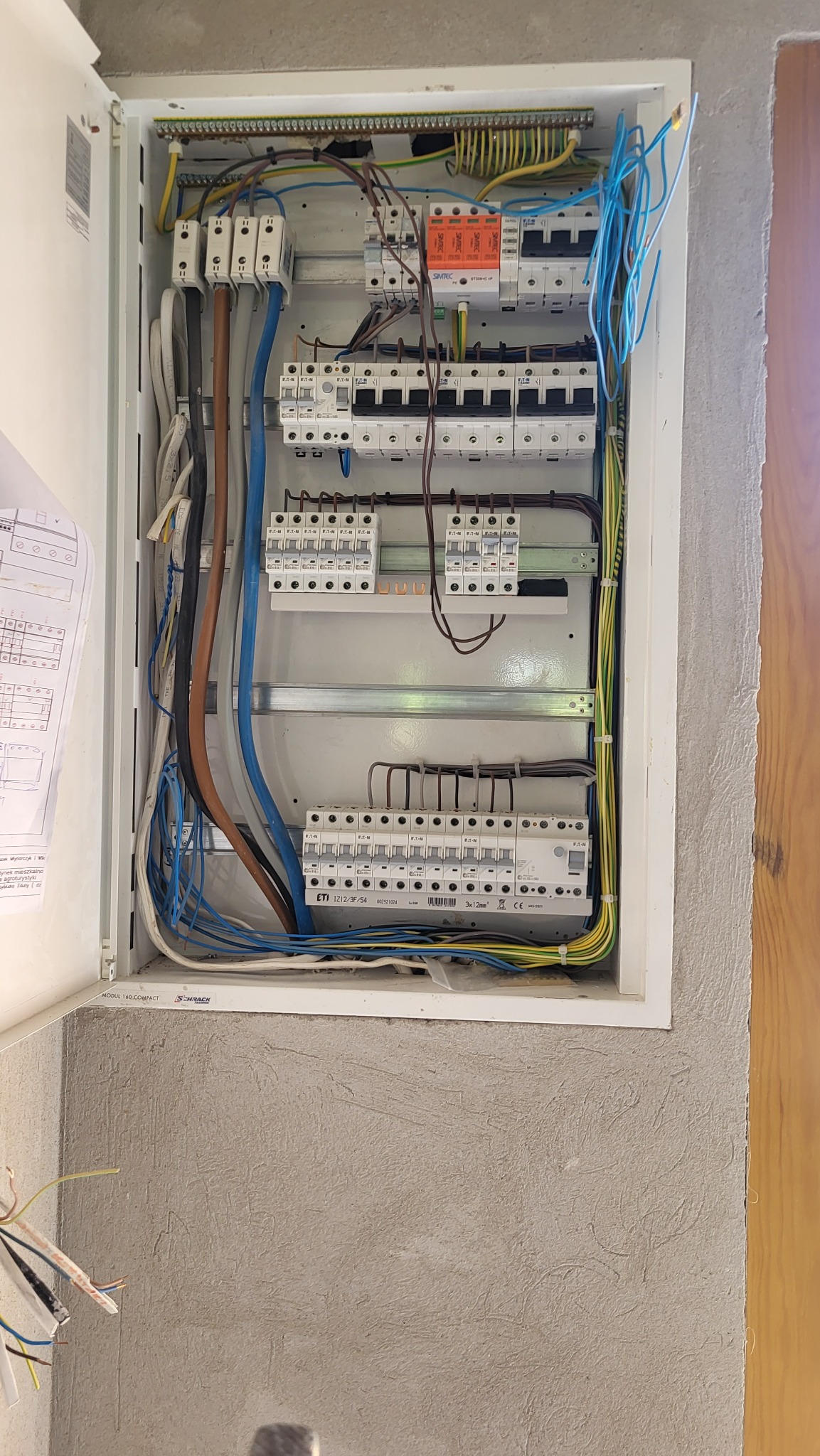 Otwarta skrzynka elektryczna z bezpiecznikami, przewodami w różnych kolorach, listwą zaciskową, zamontowana na szarej, chropowatej ścianie, widoczny schemat instalacji po lewej stronie.