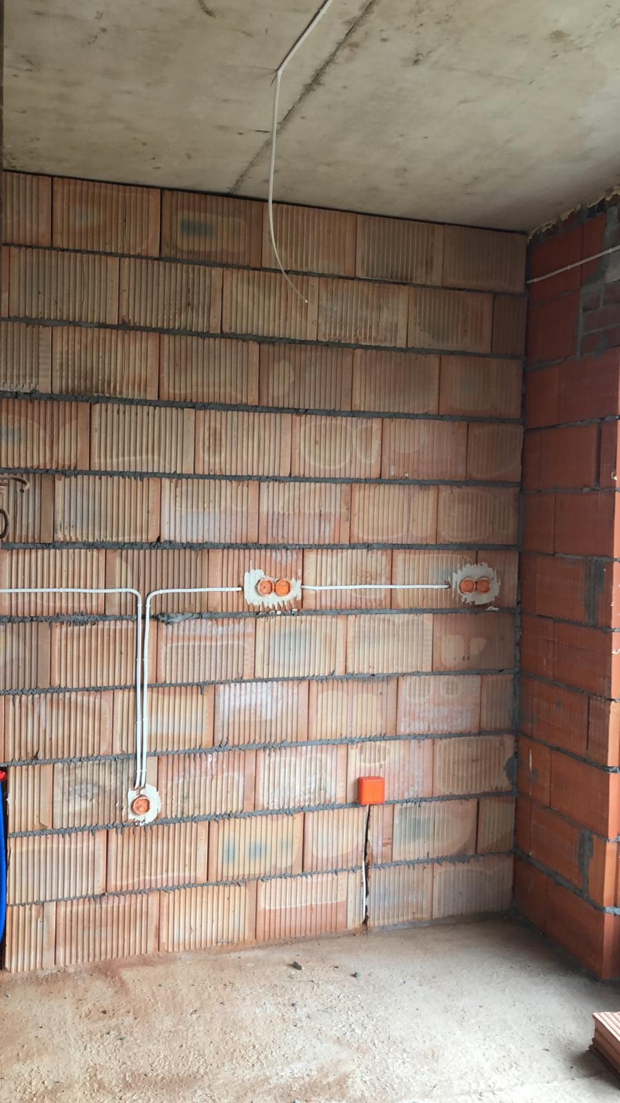 Surowa ściana z cegły ceramicznej z widocznymi przewodami elektrycznymi w białych rurkach i pomarańczowymi puszkami pod gniazdka, sufit betonowy w tle.