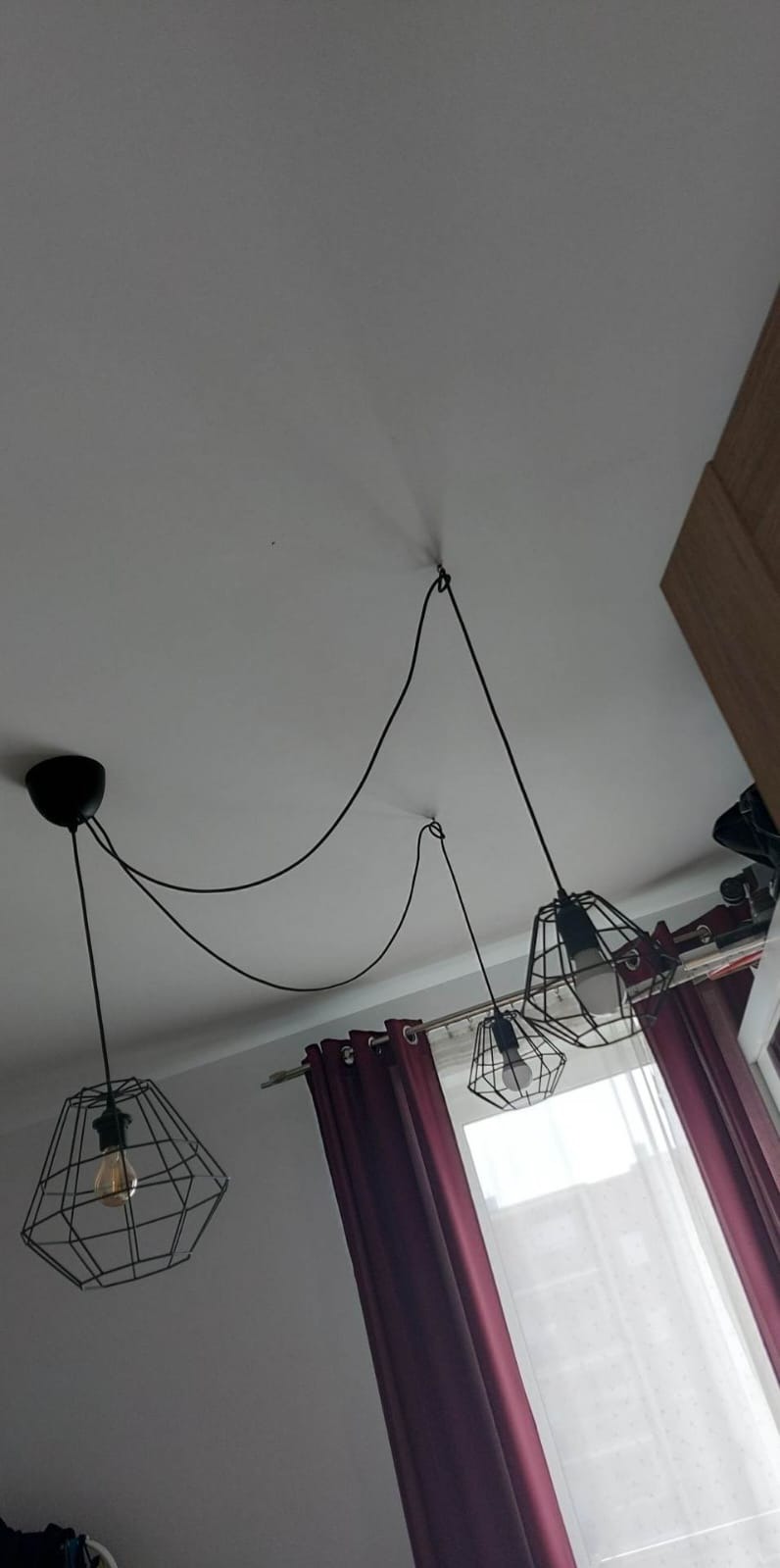 Trzy czarne, geometryczne lampy wiszące na długich kablach, zamocowane do sufitu w pokoju z bordowymi zasłonami i białym oknem, widok z dołu.