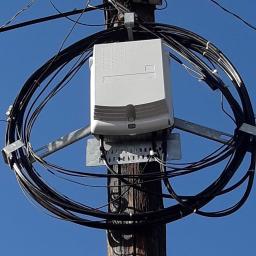 Inter-top - Skrzynka telekomunikacyjna z kablami zamontowana na drewnianym słupie na tle błękitnego nieba.