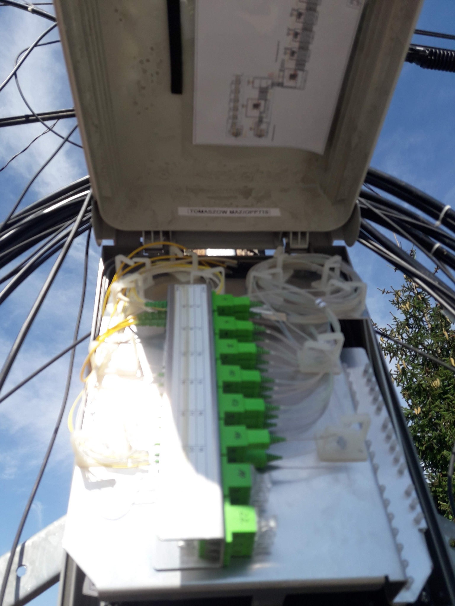 Otwarta skrzynka telekomunikacyjna z widocznymi kablami światłowodowymi, złączami i schematem, na tle błękitnego nieba i zieleni drzew.