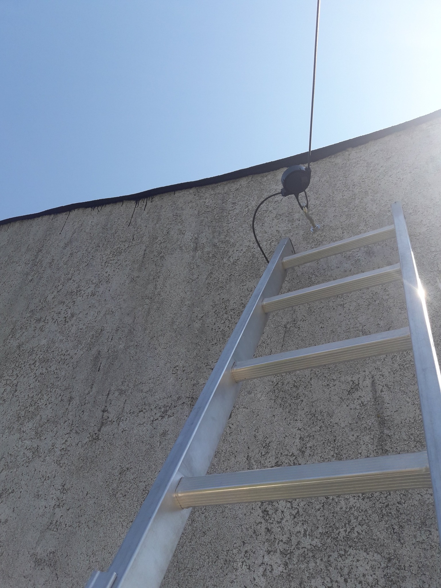 Aluminiowa drabina oparta o szorstką ścianę budynku, z zamontowaną puszką telekomunikacyjną i kablem antenowym pod błękitnym niebem.