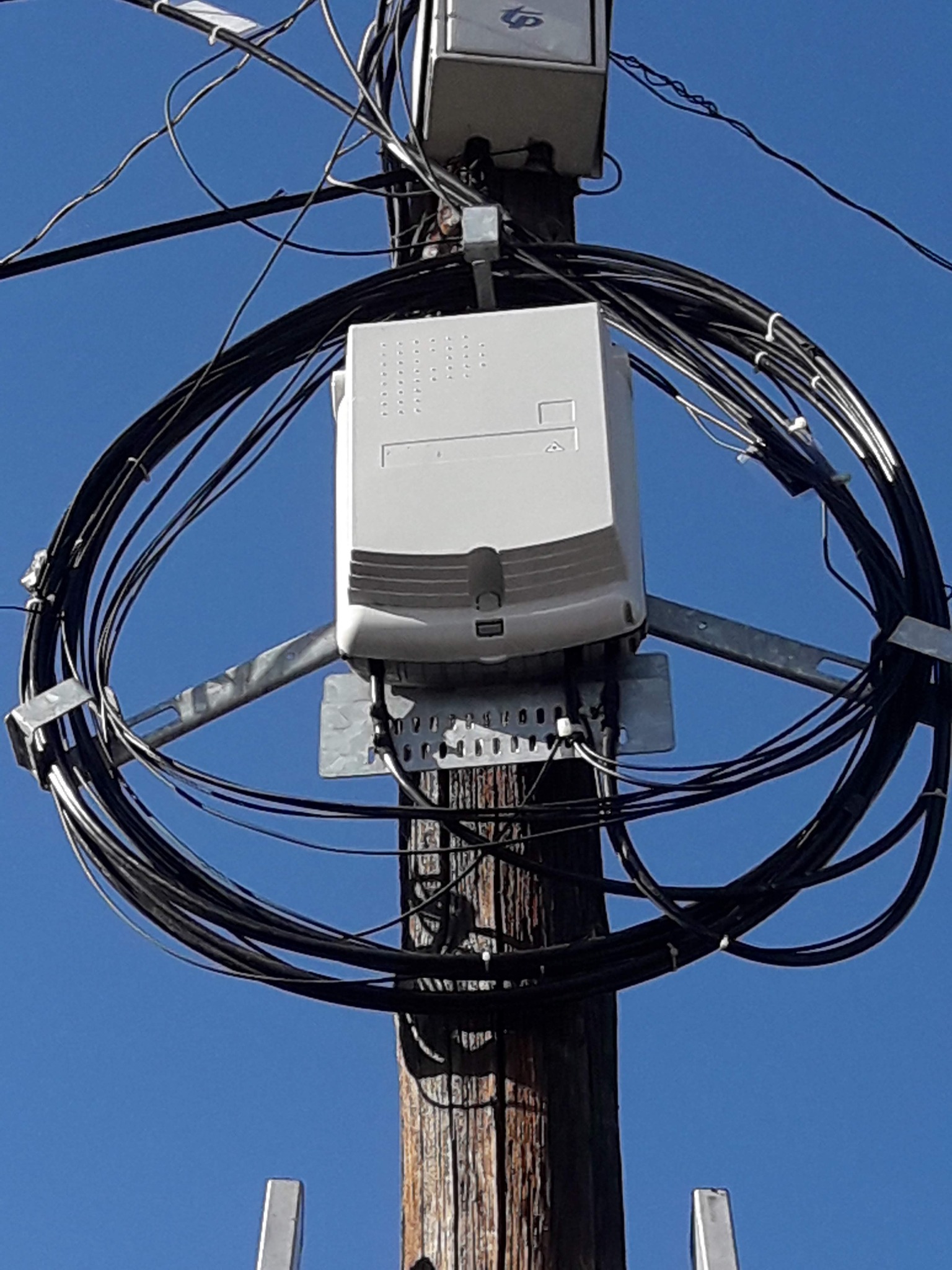 Skrzynka telekomunikacyjna z kablami zamontowana na drewnianym słupie na tle błękitnego nieba.