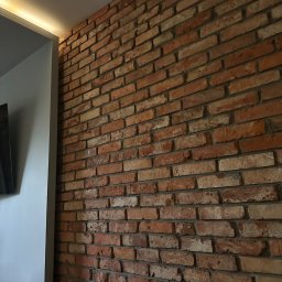 Piotr Baniewicz - Dekoracyjna ściana z cegły w salonie z podświetleniem LED, tworząca ciepły i nowoczesny klimat wnętrza. Widoczny fragment telewizora.