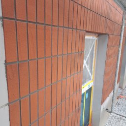 Piotr Baniewicz - Elewacja budynku z cegły klinkierowej w trakcie prac wykończeniowych, widoczne rusztowanie i zabezpieczone okno. Detal elewacji z bliska, cegła w odcieniach czerwieni.