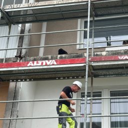 Piotr Baniewicz - Pracownik w kasku na rusztowaniu przy budynku. Widoczne uszkodzenia balkonu i elewacji. Prace remontowe w toku, z bliska, w dzień.