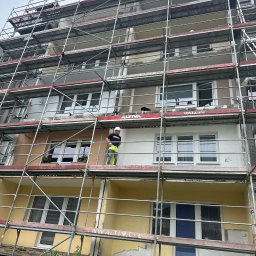 Piotr Baniewicz - Pracownik w kasku na rusztowaniu maluje elewację bloku. Widoczne fragmenty odnowionej i starej fasady. Ujęcie z dołu, na pierwszym planie zieleń.