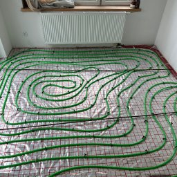 Piotr Baniewicz - Instalacja ogrzewania podłogowego: zielone rurki ułożone spiralnie na srebrnej folii z czerwoną siatką, widok z góry, grzejnik na ścianie, okno w tle.
