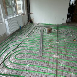 Piotr Baniewicz - Zielone rury ogrzewania podłogowego ułożone na siatce w remontowanym pomieszczeniu, widoczny grzejnik, okno i wiadro z zaprawą.