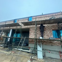 Piotr Baniewicz - Budowa domu z cegły: murarze na rusztowaniu układają cegły, okna zabezpieczone folią. Widok z dołu na budynek w trakcie wznoszenia.