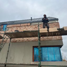 Piotr Baniewicz - Mężczyzna na rusztowaniu podczas prac murarskich na elewacji budynku z cegły klinkierowej. Okna zabezpieczone folią. Widoczne narzędzia i materiały budowlane.