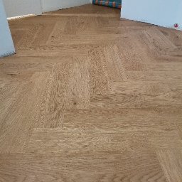 Garant-BUD - Ułożona w jodełkę jasna drewniana podłoga w nowym pomieszczeniu z białymi ścianami, zabezpieczonymi folią fragmentami ścian i widocznymi gniazdkami elektrycznymi.