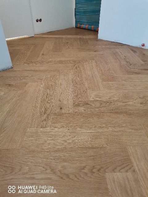 Ułożona w jodełkę jasna drewniana podłoga w nowym pomieszczeniu z białymi ścianami, zabezpieczonymi folią fragmentami ścian i widocznymi gniazdkami elektrycznymi.