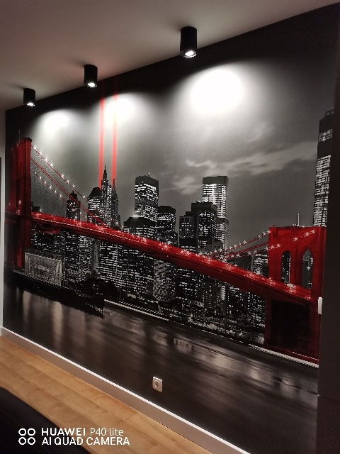 Fototapeta z motywem panoramy Nowego Jorku z czerwonym mostem, oświetlona punktowymi lampami sufitowymi, z widoczną listwą przypodłogową i gniazdkiem elektrycznym.