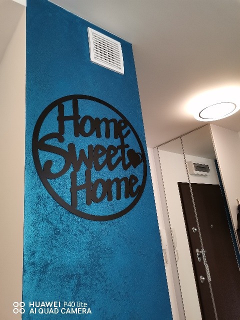 Błękitna ściana z dekoracyjną strukturą i czarnym napisem 'Home Sweet Home', wentylacja w górnej części ściany, widoczny fragment przedpokoju z drzwiami.