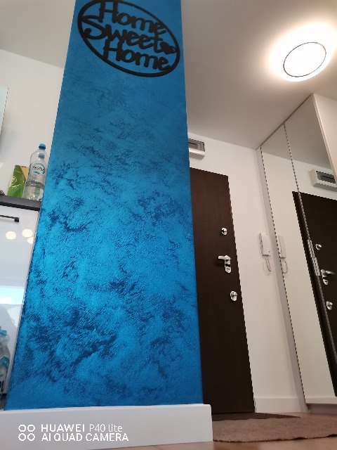 Pionowy słup w przedpokoju pokryty dekoracyjną, turkusową farbą z efektem marmuru, ozdobiony czarnym napisem 'Home Sweet Home', widoczne drzwi wejściowe i oświetlenie sufitowe.