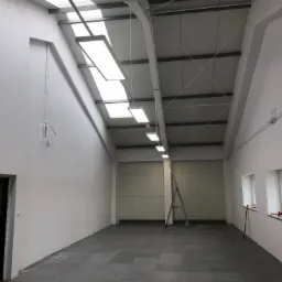 Wnętrze hali przemysłowej z zainstalowanym oświetleniem LED, widoczne okna dachowe, drabina i instalacja elektryczna na ścianie.
