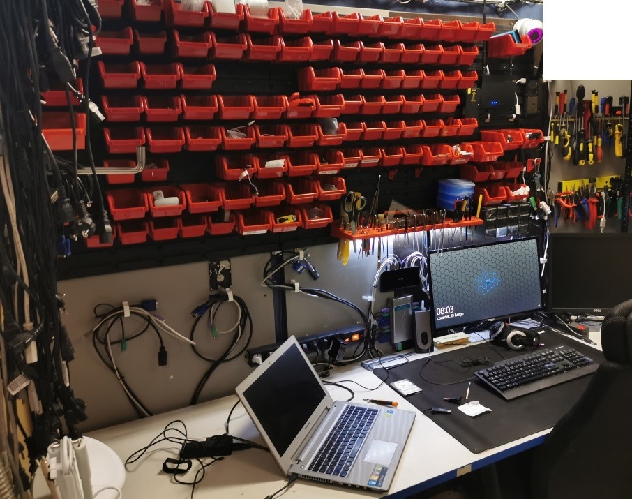 Stanowisko pracy serwisanta komputerowego z laptopem, monitorem, narzędziami i ścianą pełną czerwonych pojemników na części zamienne.