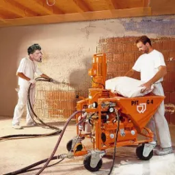 Dwóch pracowników podczas nakładania tynku gipsowego na ścianę z cegły, używając pomarańczowej maszyny PFT G4 z podłączonymi wężami.