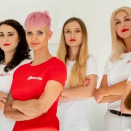 Grupa sześciu kobiet, w tym jedna z krótkimi różowymi włosami i w czerwonej koszulce, stojąca w gabinecie z białymi ścianami i szafkami, wszystkie z założonymi rękami, prezentujące zespół.