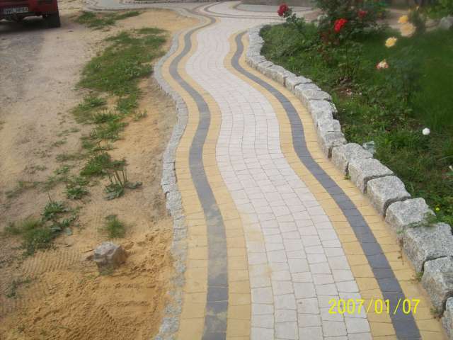 Kręta ścieżka z kostki brukowej w kolorach żółtym, szarym i białym, obrzeża z kamieni granitowych, widok z góry, częściowo zarośnięta trawą, w tle czerwony samochód.