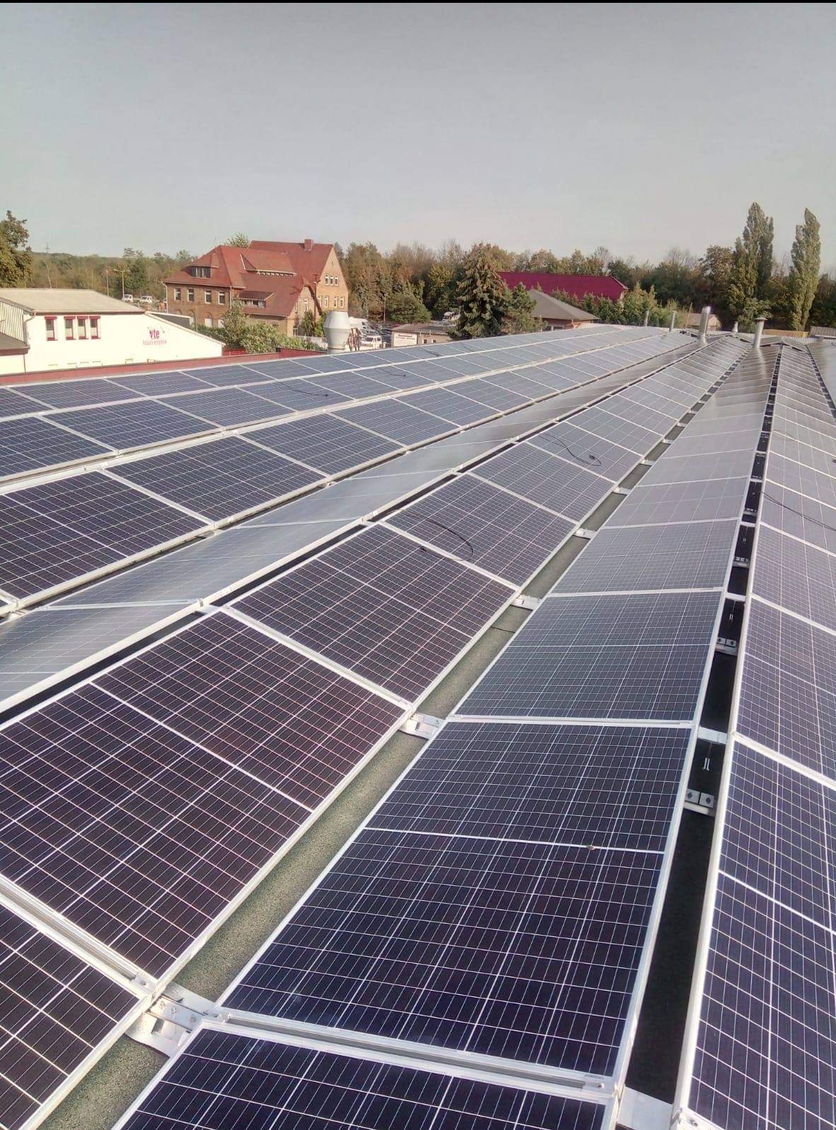 Rozległa instalacja fotowoltaiczna na dachu budynku przemysłowego, widok z góry, w tle budynki i drzewa.