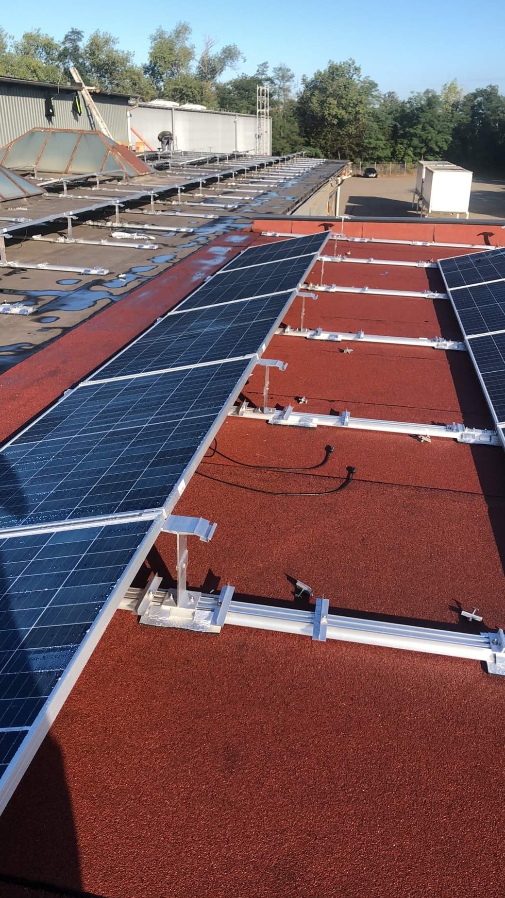 Instalacja paneli słonecznych na dachu pokrytym czerwoną nawierzchnią, widoczne aluminiowe konstrukcje montażowe i kable.