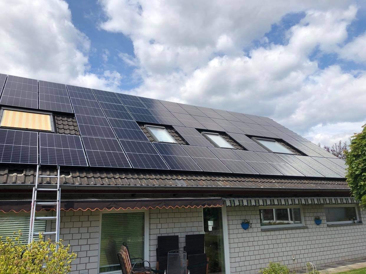 Instalacja paneli słonecznych na dachu domu jednorodzinnego z oknami dachowymi i rozłożoną markizą nad tarasem, drabina oparta o dach.