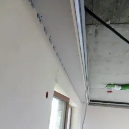 Montaż płyty gipsowo-kartonowej na suficie podwieszanym, widoczne profile metalowe i taśma mocująca Rigips, fragment okna i instalacja wentylacyjna w tle.