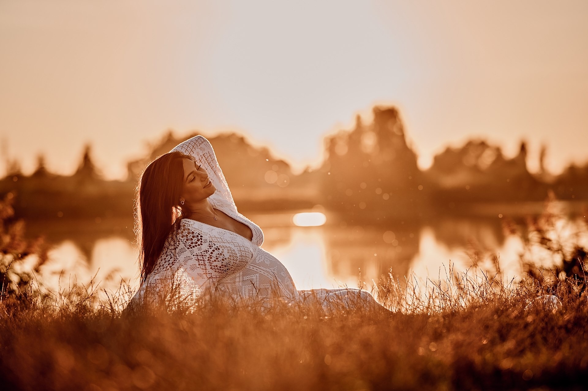 Kobieta w ciąży w koronkowej sukni leży na trawie nad brzegiem jeziora podczas zachodu słońca, z ręką uniesioną za głową i zamkniętymi oczami.