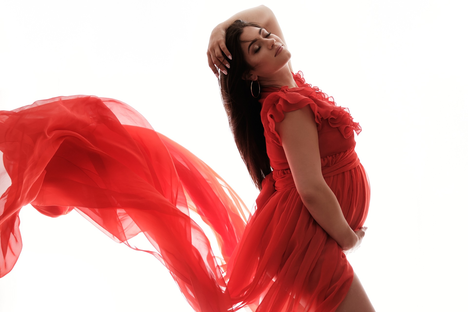 Kobieta w czerwonej sukience z falującym materiałem, dłoń na głowie, zamknięte oczy, białe tło.