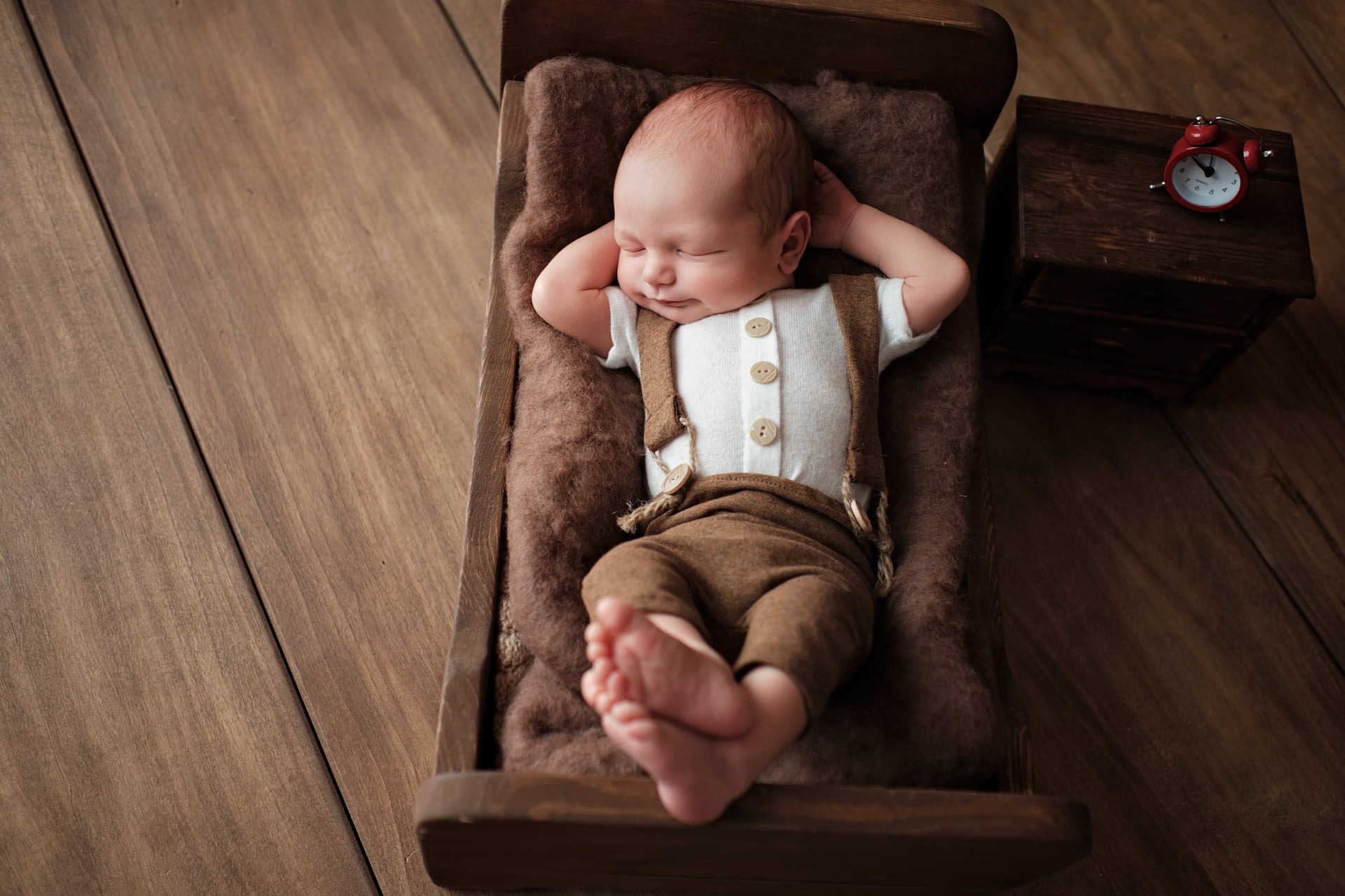 Śpiący noworodek w brązowym ubranku z szelkami, ułożony na miękkim kocyku w miniaturowym drewnianym łóżeczku obok szafki nocnej z czerwonym budzikiem.