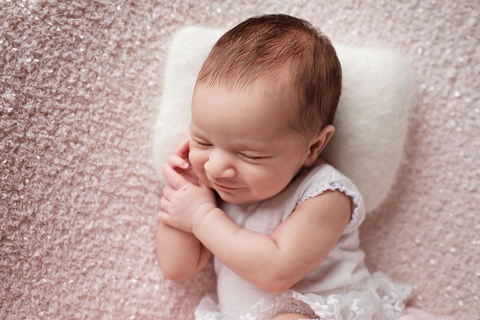 Śpiąca noworodka ułożona na białej poduszce na tle teksturowanego, różowego koca. Rączki dziecka złożone blisko twarzy.