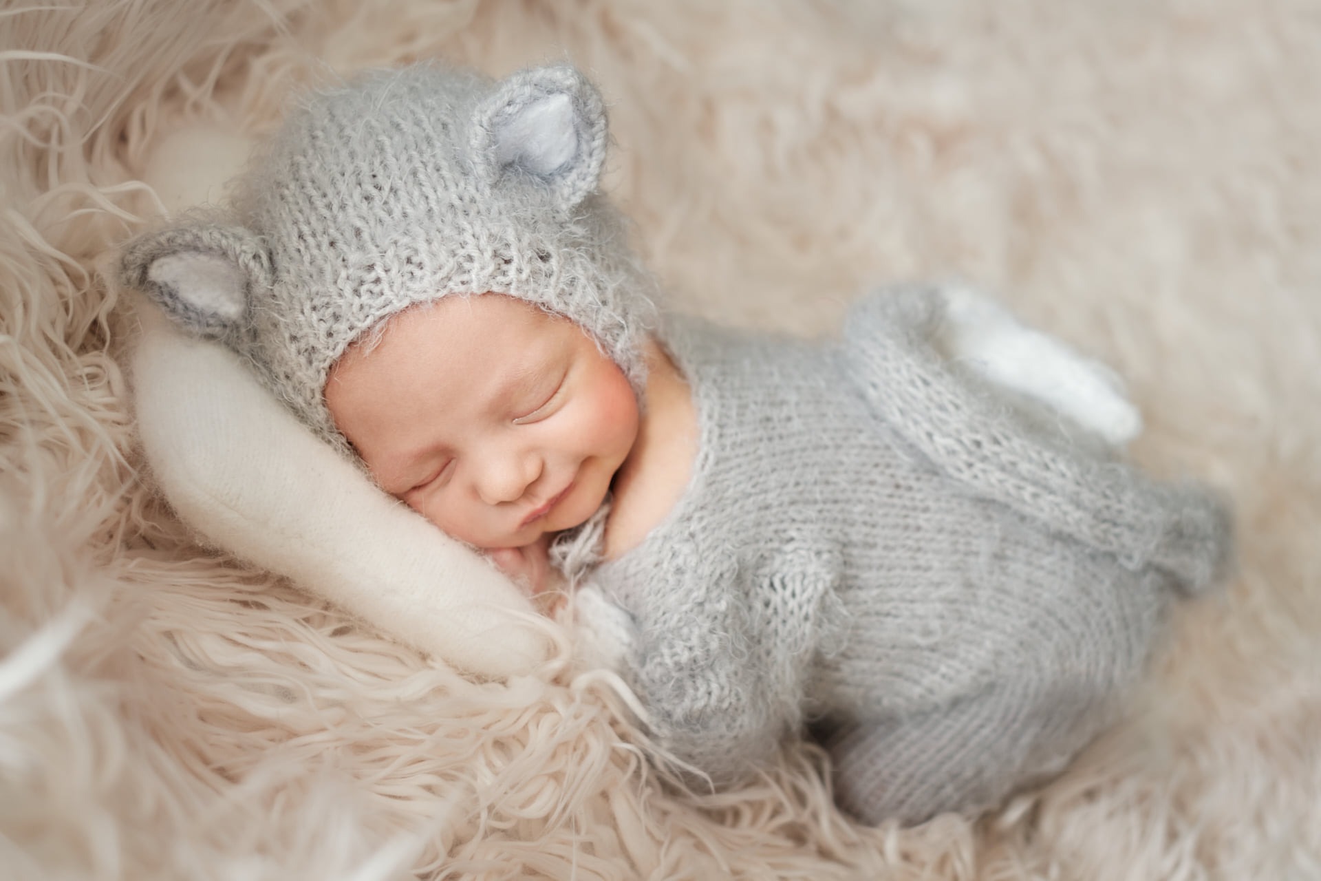 Śpiący noworodek w szarym, dzianinowym kombinezonie z uszkami, leżący na puszystym, kremowym kocyku i białej poduszce.
