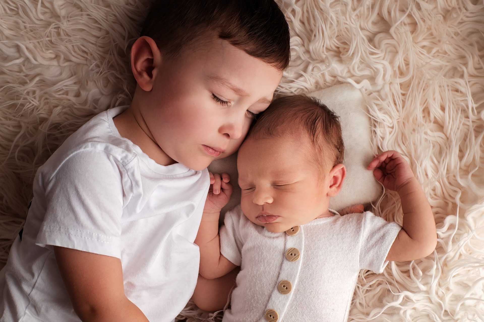 Śpiący noworodek w białym ubranku z drewnianymi guzikami, leżący obok starszego brata na puszystym, kremowym kocyku.
