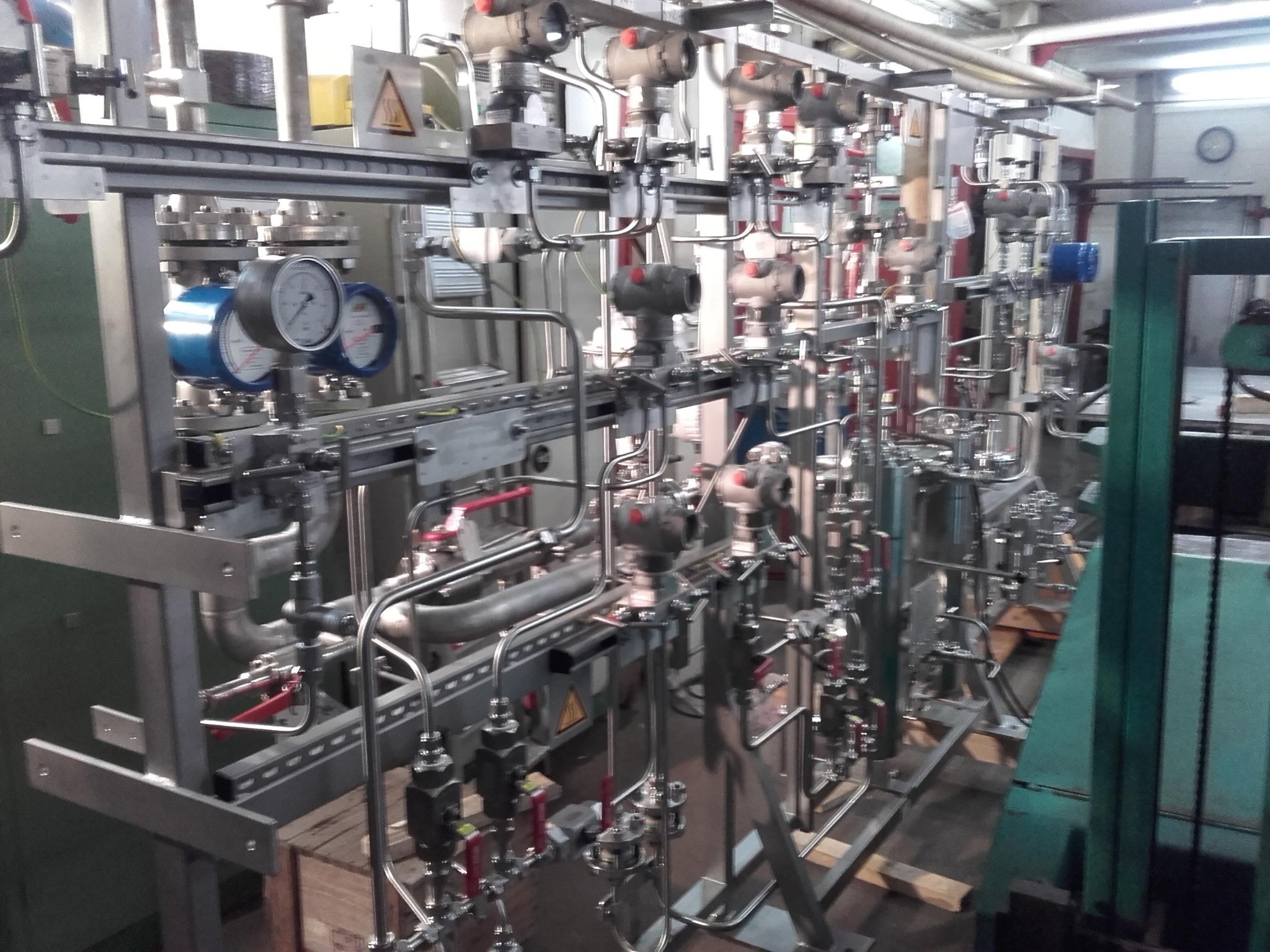 Złożona instalacja przemysłowa z rur, zaworów i manometrów, widoczna tabliczka ostrzegawcza z trójkątem, w tle fragment ściany i zegar.