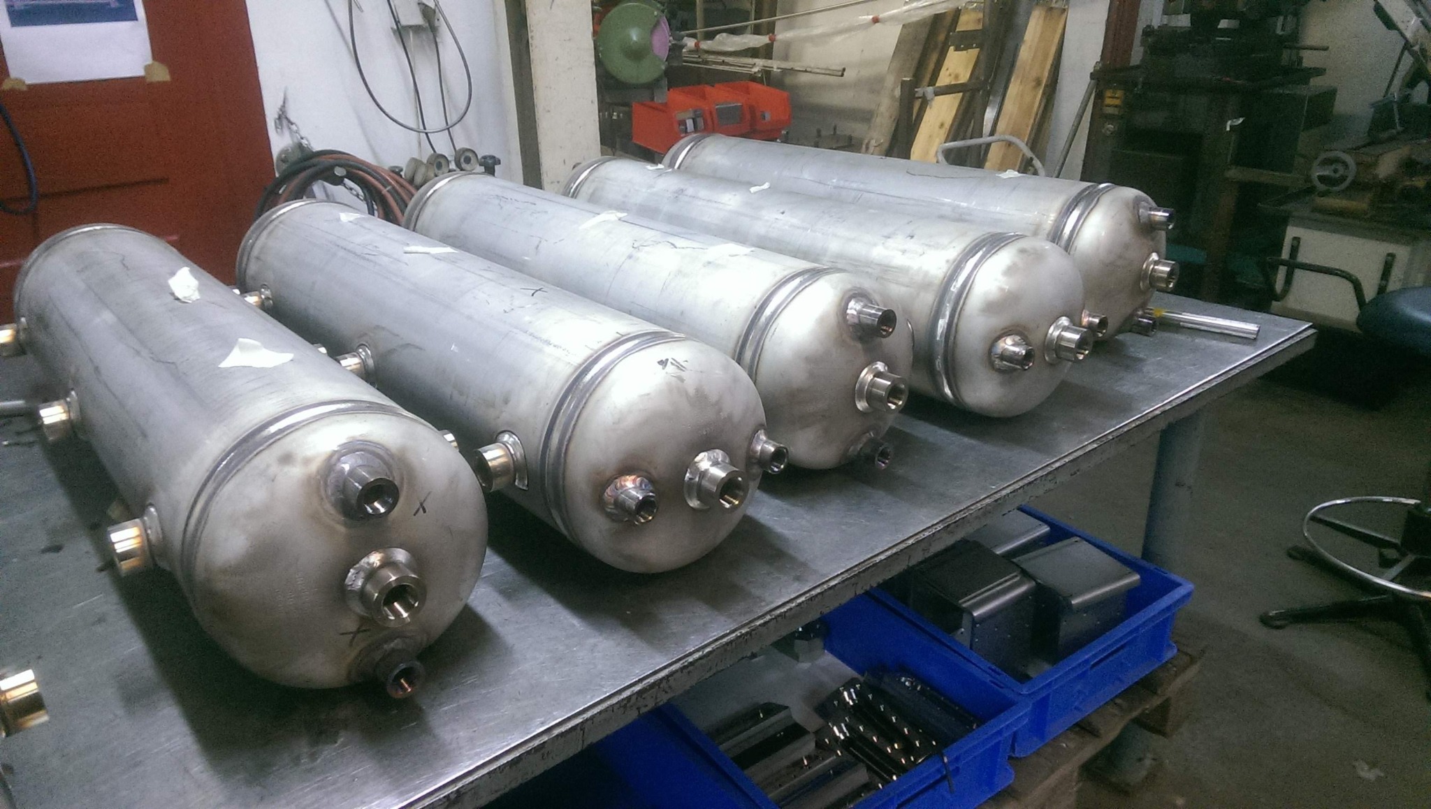 Rząd cylindrycznych zbiorników ze stali nierdzewnej, każdy z trzema króćcami, ułożonych na metalowym stole w warsztacie.