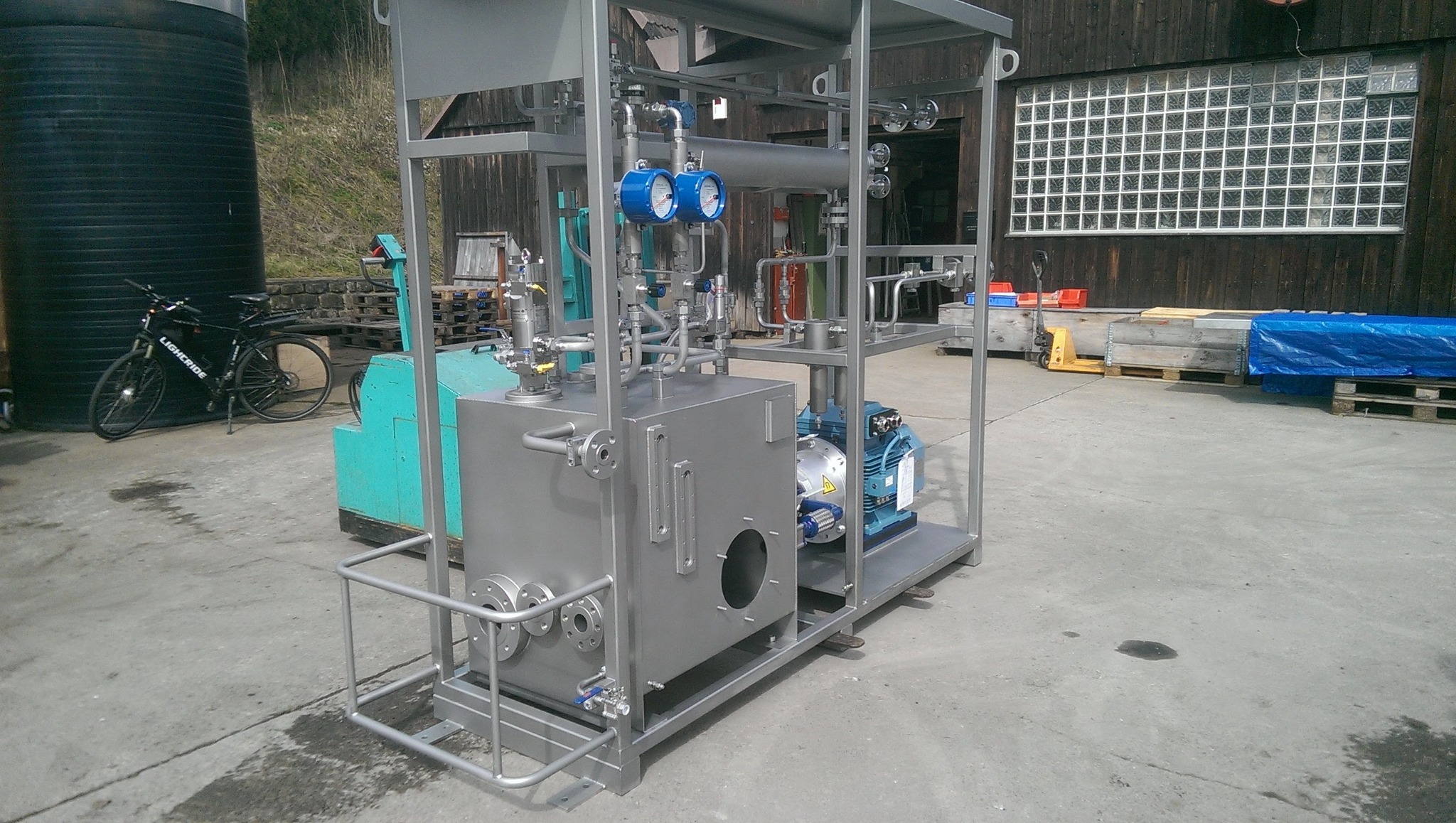 Stalowa konstrukcja z zbiornikiem, rurami, manometrami i silnikiem elektrycznym, umieszczona na zewnątrz, widoczny rower i budynek w tle.