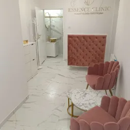 Wnętrze kliniki kosmetycznej z marmurową podłogą, różowymi fotelami w stylu retro, stolikiem z marmurowym blatem i recepcją z pikowanym frontem, widok na łazienkę z elementami złota.