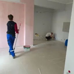Malowanie ścian na różowo wałkiem, drugi pracownik szpachluje narożnik w pomieszczeniu z szarymi płytkami podłogowymi i niezabezpieczonymi kablami.