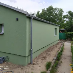 Świeżo pomalowana elewacja budynku na zielono, widoczne okna, rynna, lampa zewnętrzna i ścieżka wzdłuż ściany.