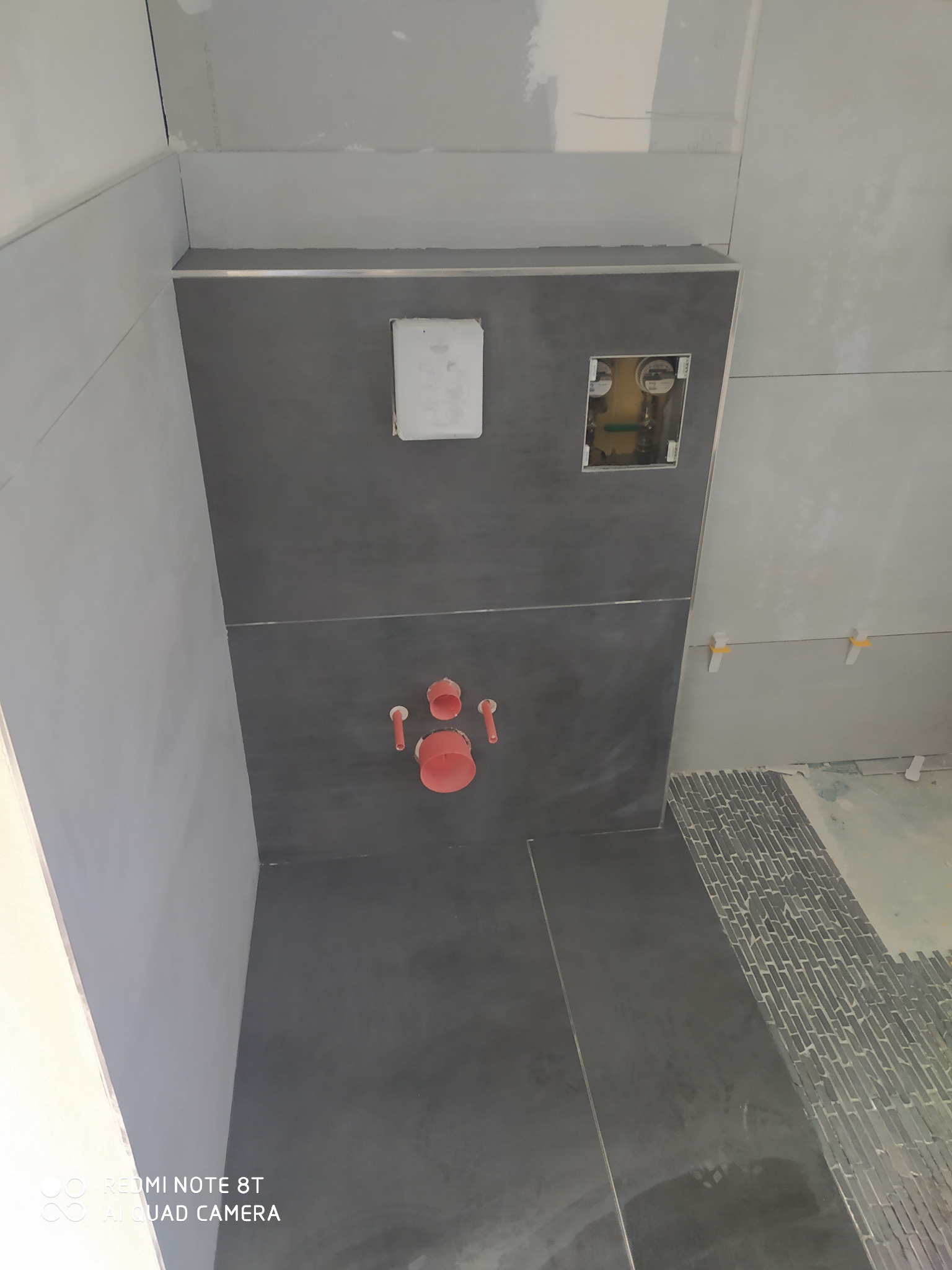 Montaż podtynkowego stelaża WC z szarą okładziną ścienną i fragmentem ułożonej mozaiki podłogowej w trakcie remontu łazienki.