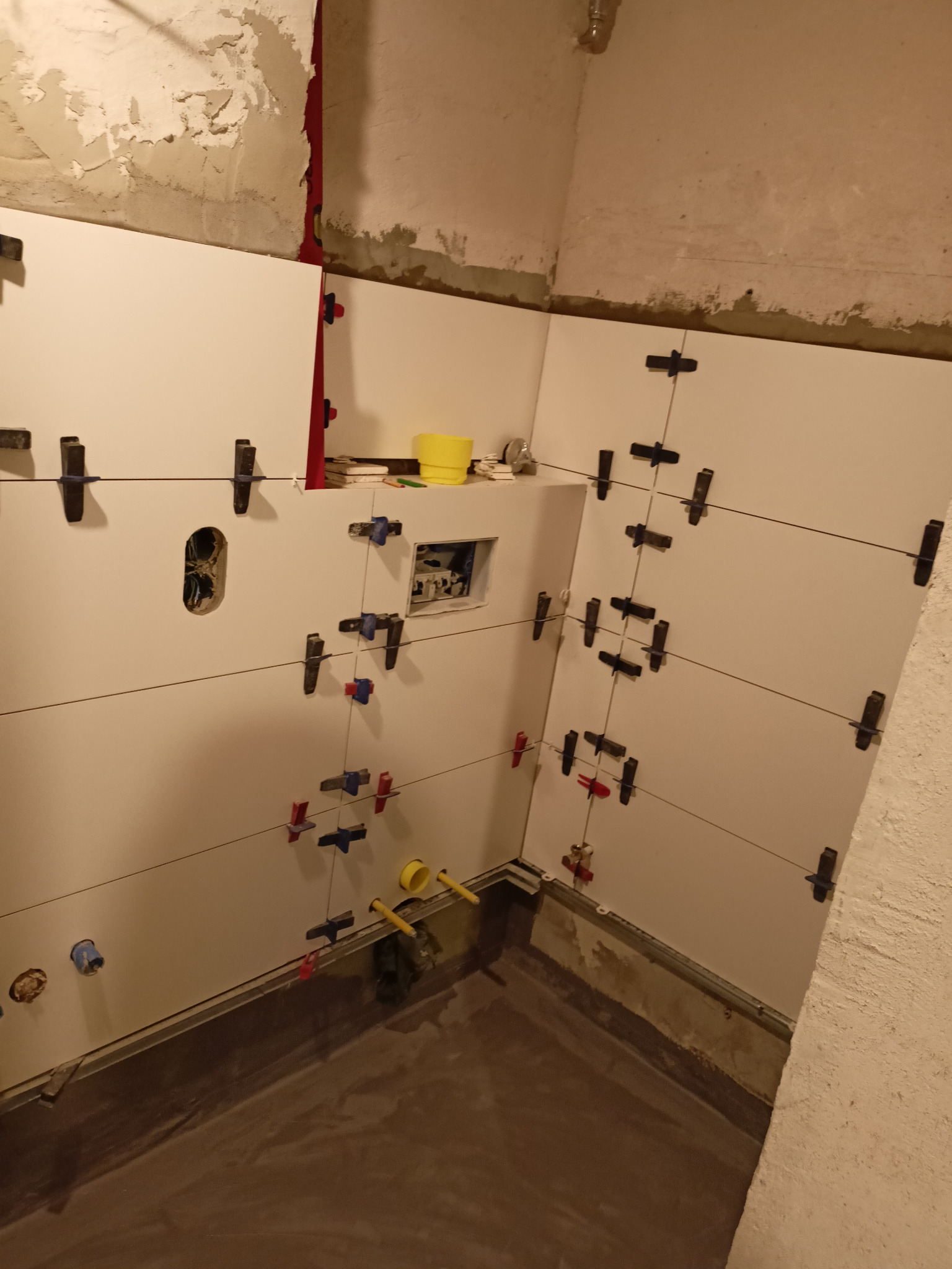 Świeżo ułożone, białe płytki ścienne w trakcie fugowania, z użyciem krzyżyków dystansowych i systemu poziomowania, widoczne instalacje hydrauliczne i elektryczne, surowy stan ścian i podłogi.