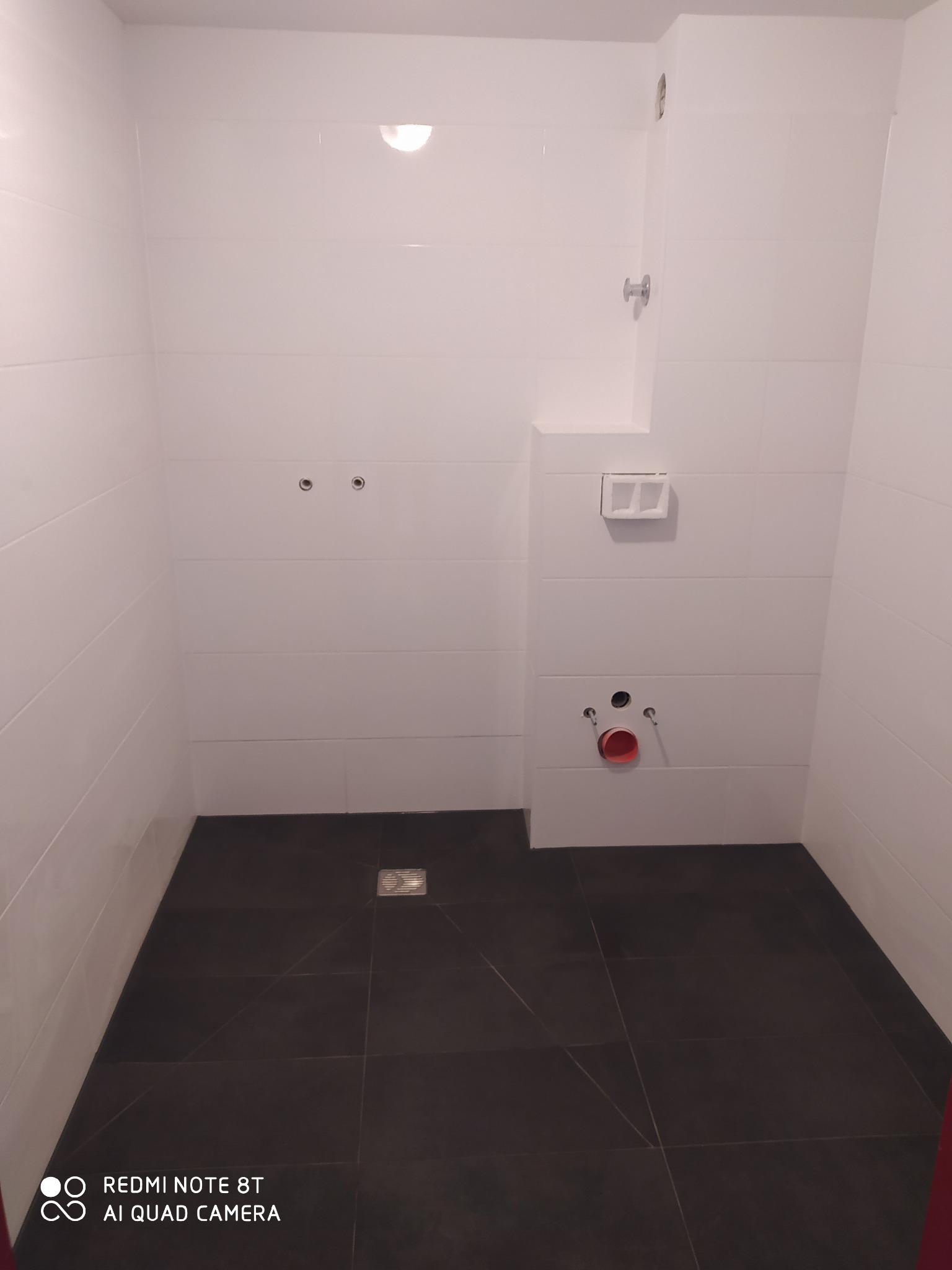 Pusta łazienka w trakcie prac wykończeniowych: białe płytki ścienne, ciemna podłoga z odpływem liniowym, przygotowane przyłącza wodno-kanalizacyjne.